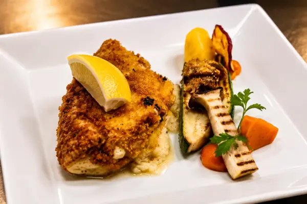 Goldbraun gebackenes Cordon bleu vom Schwein mit Zitronenschnitz, gegrillten Kräuterseitlingen, Zucchini und Karotte auf weissem Teller im Gasthof Kreuz Egerkingen