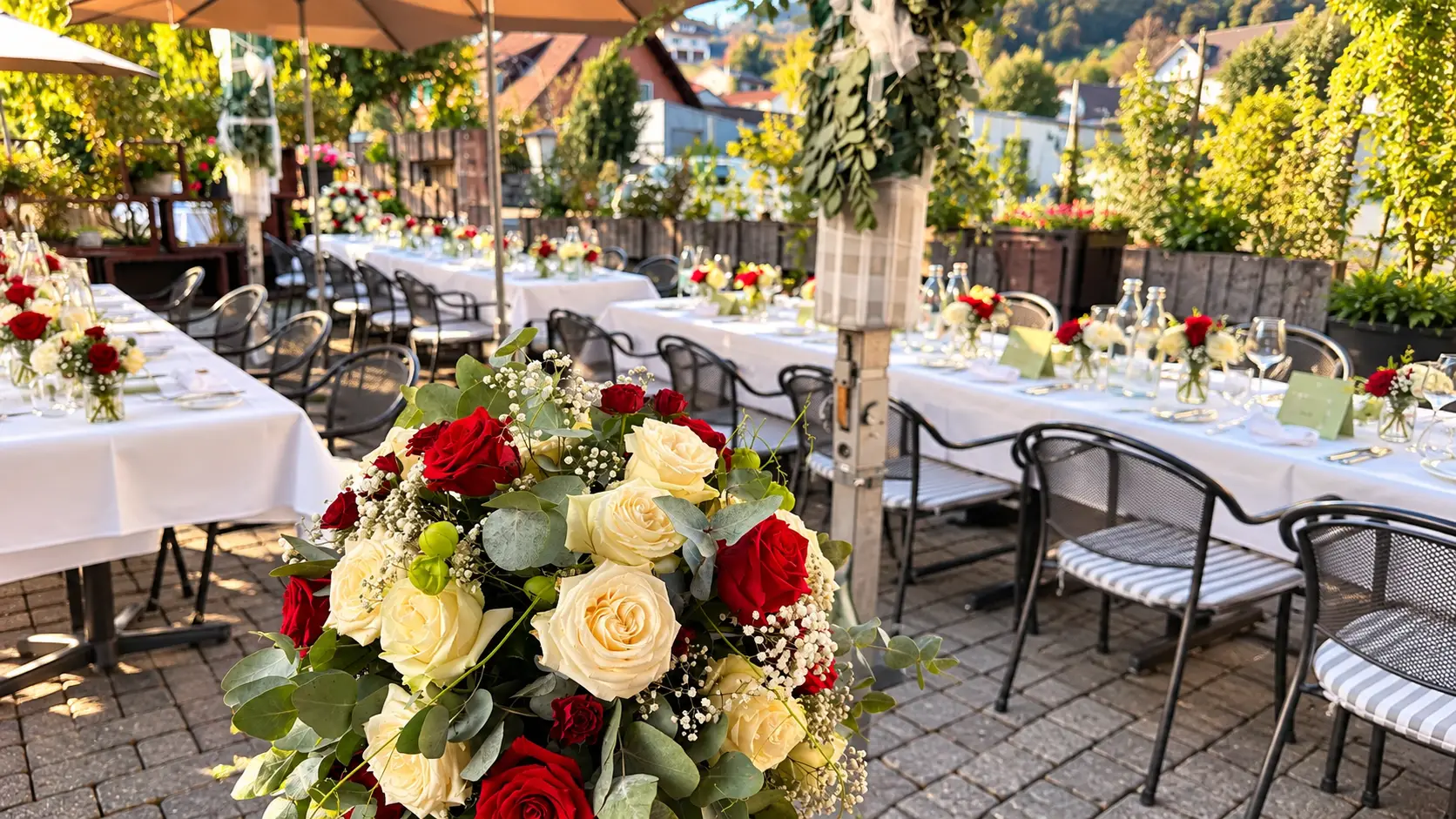Grosses Gesteck aus roten und cremefarbenen Rosen vor den gedeckten Hochzeitstischen auf der Terrasse